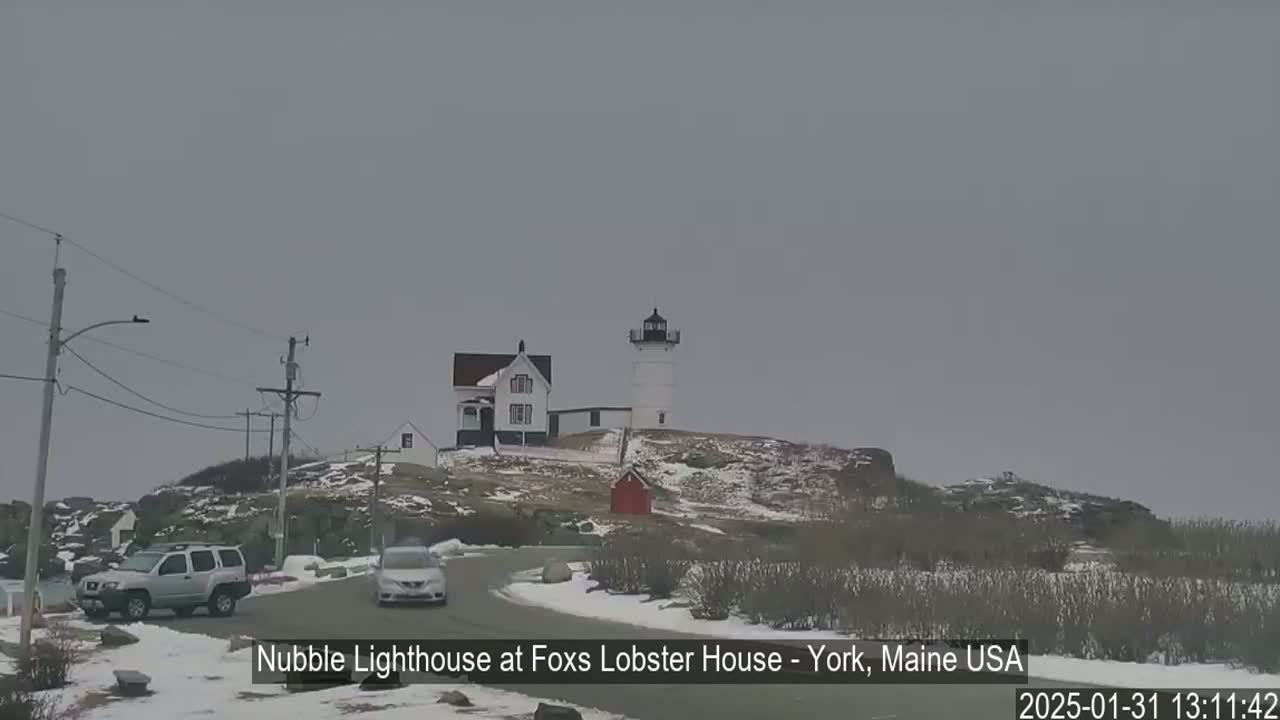A lighthouse and accompanying buildings sit atop a snow-dusted hill, with a couple of vehicles on a nearby road under an overcast sky.