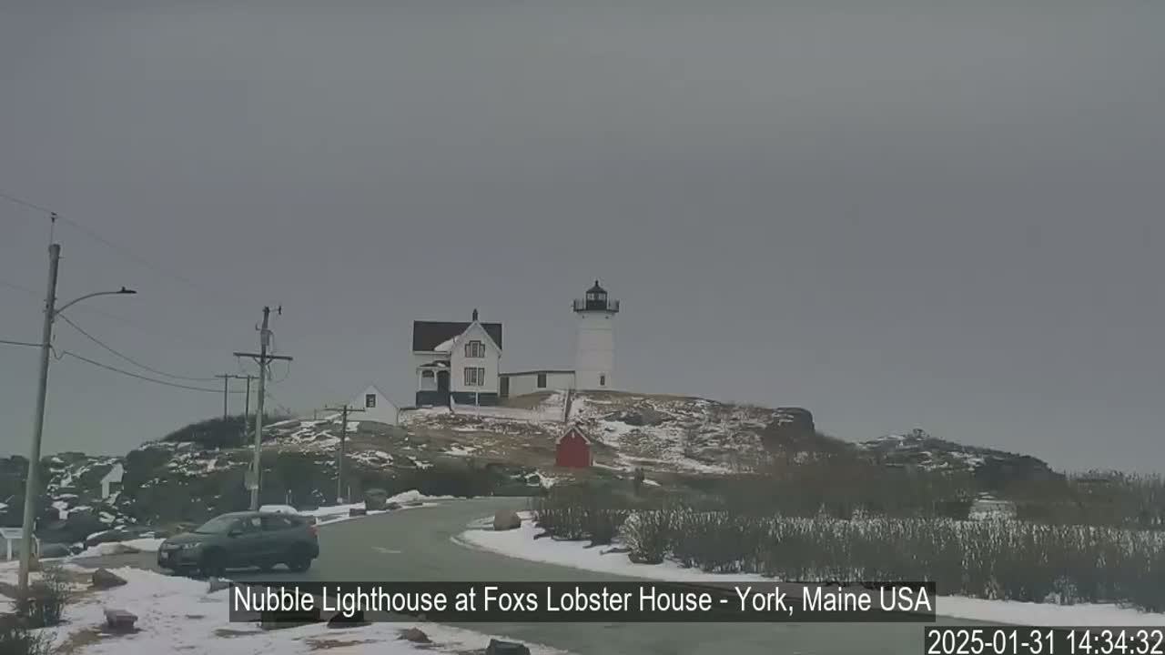 A lighthouse and keeper's house are situated on a snow-covered hill overlooking a road with a car, under an overcast sky.