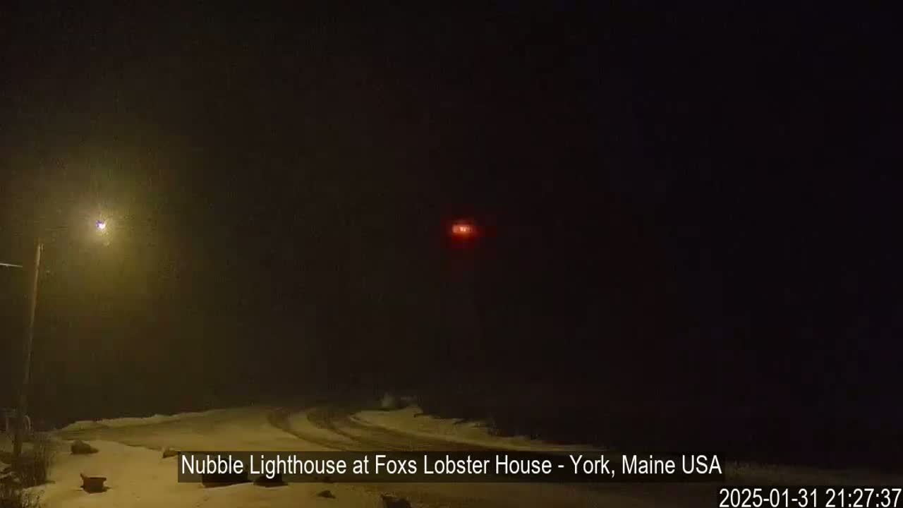 A snowy, nighttime scene shows a dimly lit area with a distant red light and a single streetlight.