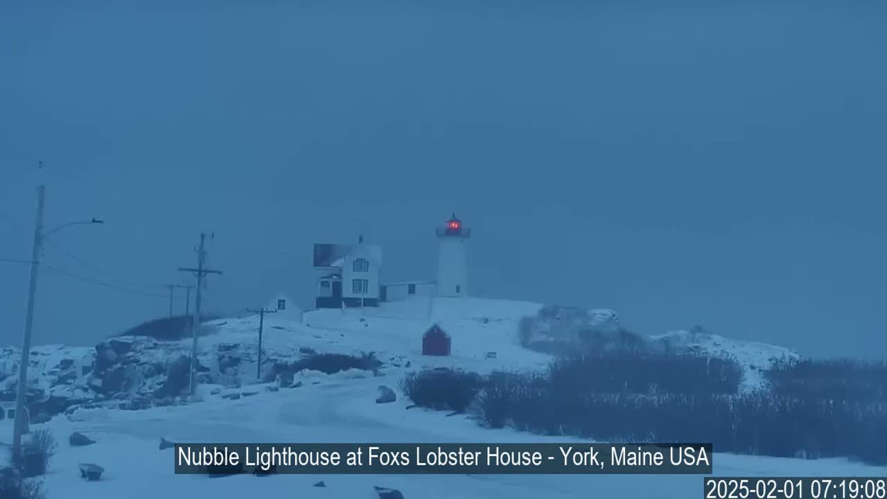 A snow-covered landscape features a lighthouse with a red light at its top, a smaller building nearby, and utility poles in the foreground under an overcast sky.