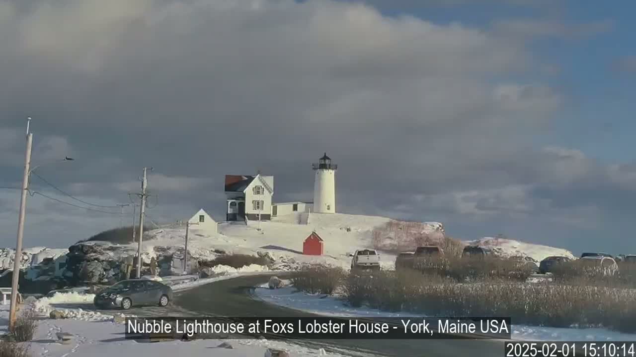 A lighthouse and accompanying buildings sit atop a snow-covered hill under a mostly cloudy sky.