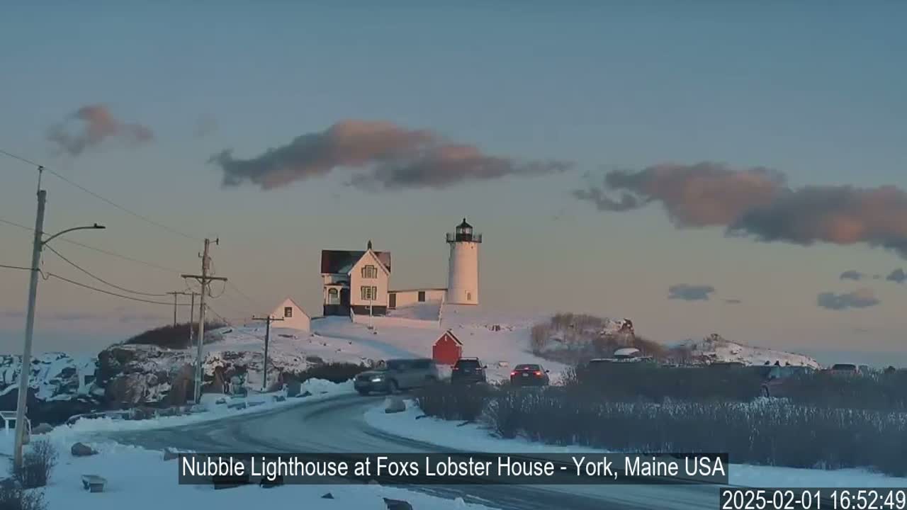 A lighthouse and accompanying buildings are situated on a snow-covered hill overlooking a winding road with several cars, under a partly cloudy twilight sky.