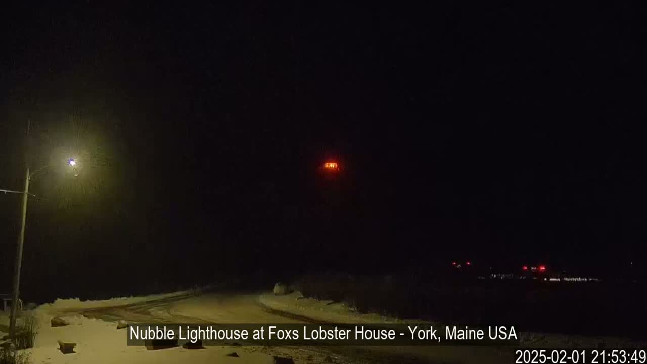 A snow-covered road at night is illuminated by a streetlight, with a distant lighthouse visible in the darkness.