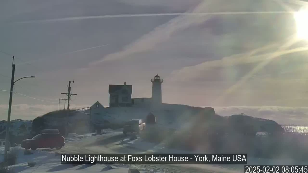 A lighthouse and other buildings sit on a snow-covered hill near a road with cars, under a partly sunny sky.