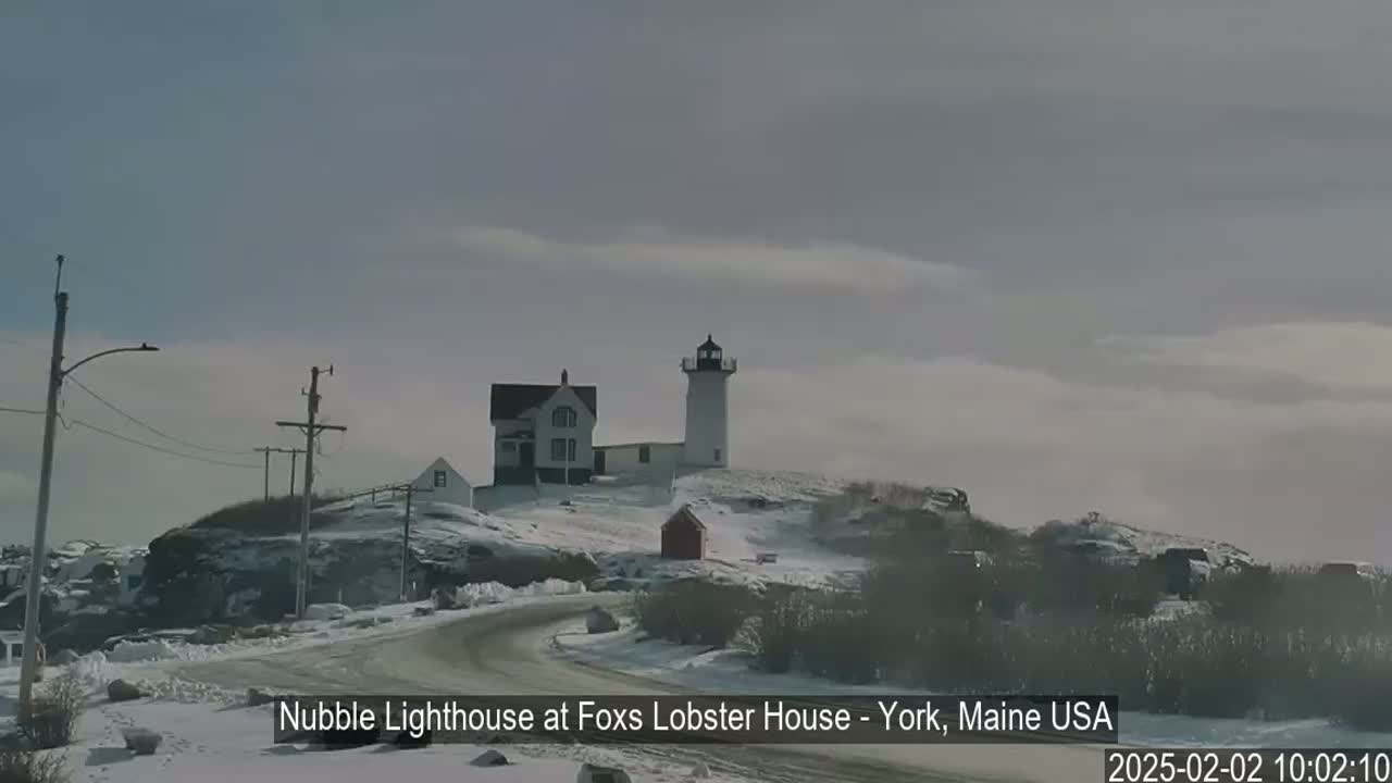 A lighthouse and a smaller building sit atop a snow-covered hill, with a winding road leading towards them under a mostly cloudy sky.