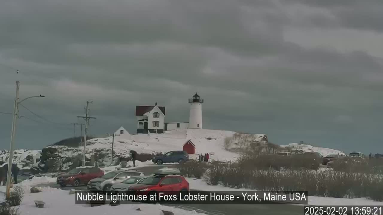A snow-covered lighthouse and keeper's house sit atop a hill, with several cars parked in a lot below under an overcast sky.