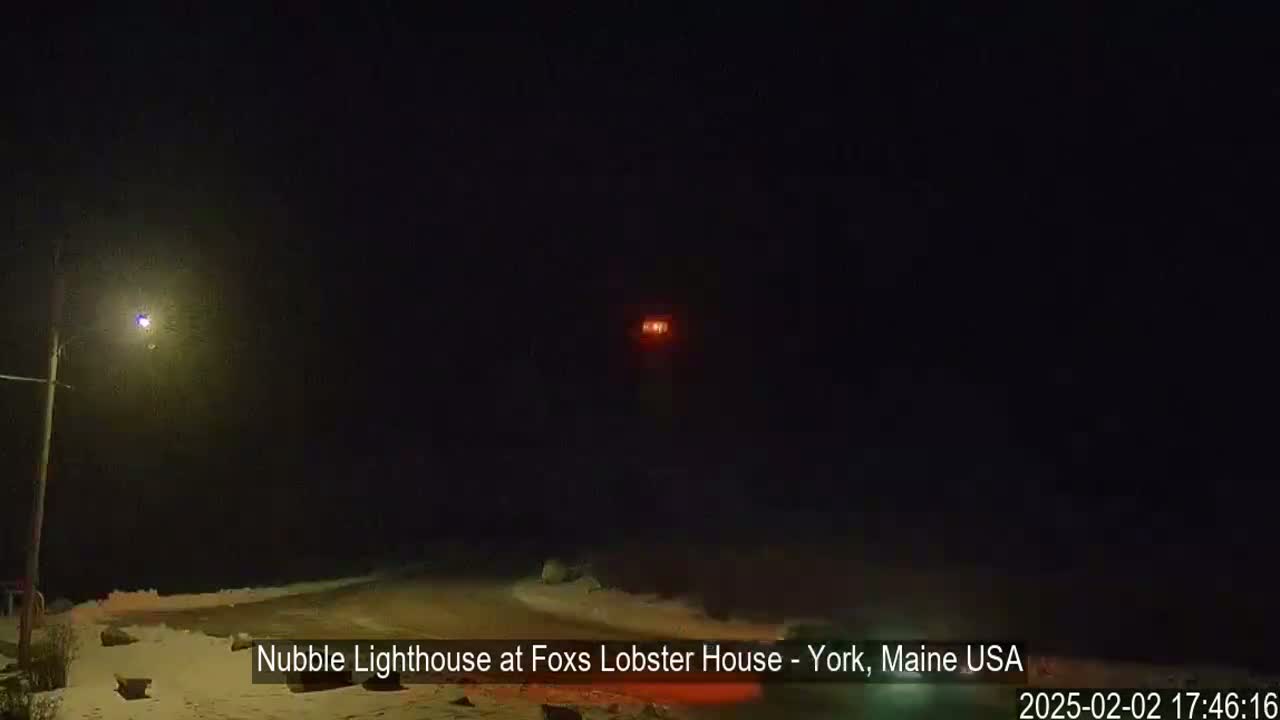 A nighttime view of a lighthouse's light, a snow-covered roadside, and a streetlight in the distance.