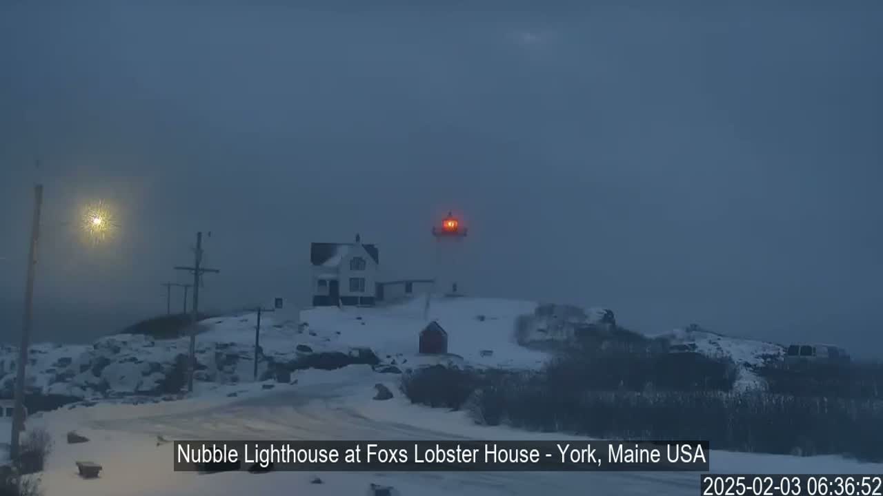 A snow-covered lighthouse with its light on is visible in foggy conditions.