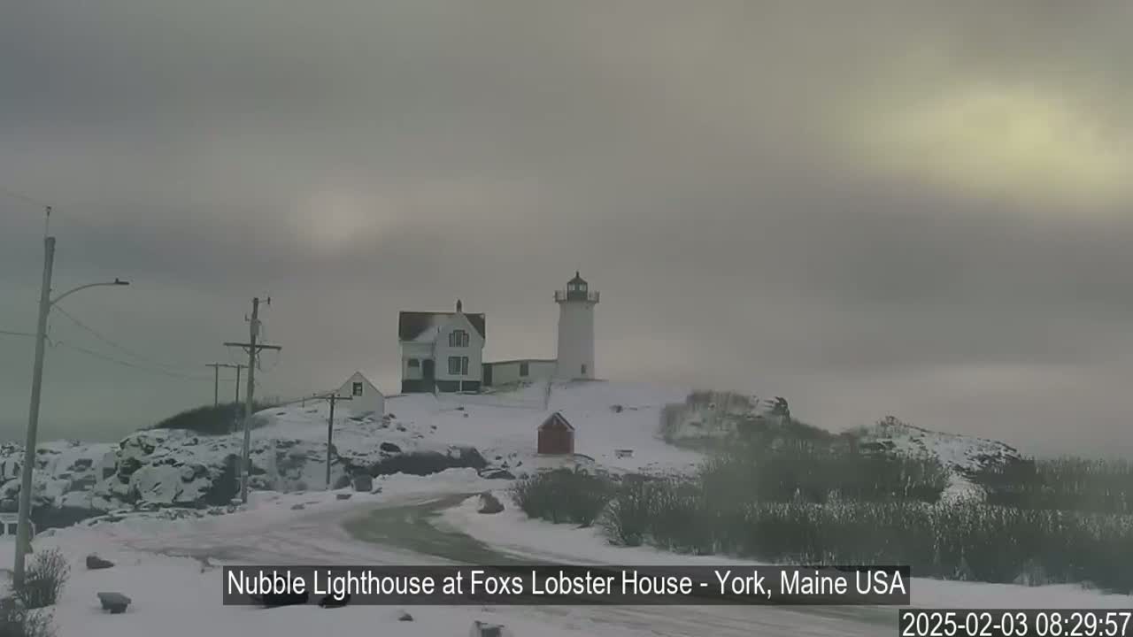 A lighthouse and buildings are situated on a snow-covered rocky outcrop under an overcast sky.