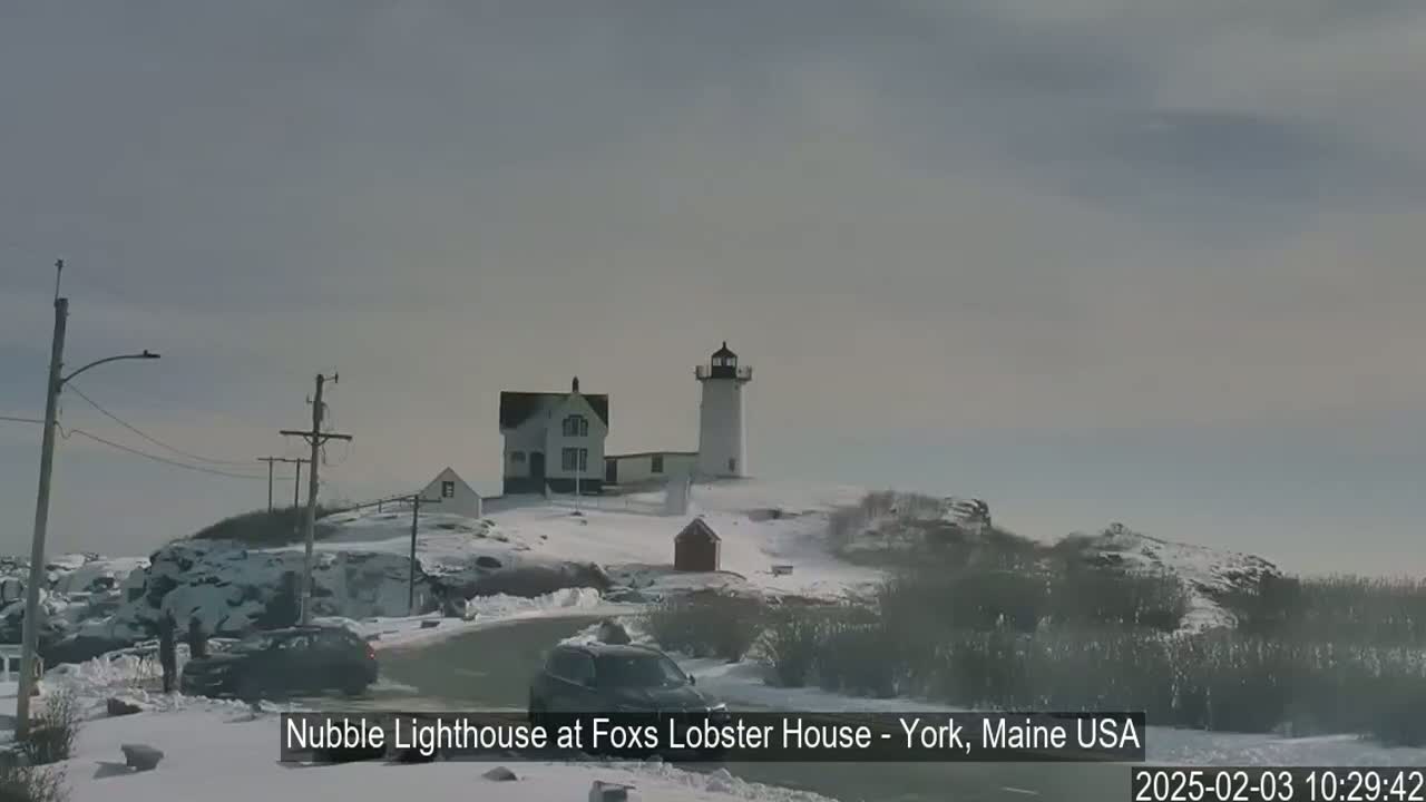 A lighthouse and a house sit atop a snow-covered hill next to a road with cars parked alongside it, under an overcast sky.