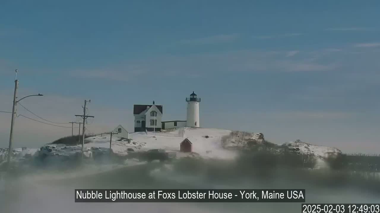 A lighthouse and keeper's house sit atop a snow-covered hill partially obscured by fog under a clear blue sky.
