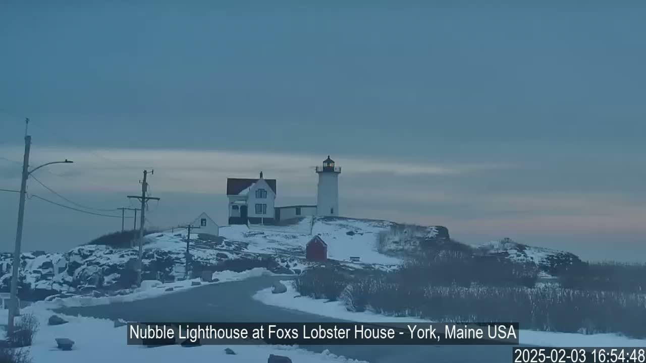 A lighthouse and other buildings are situated on a snow-covered, rocky hillside under a cloudy, twilight sky.