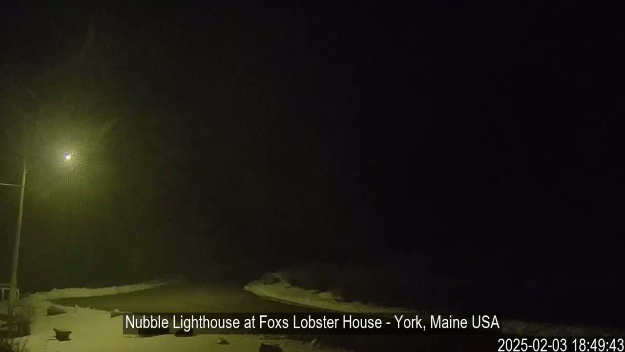 A nighttime, snowy scene shows a dimly lit road leading to a dark area.