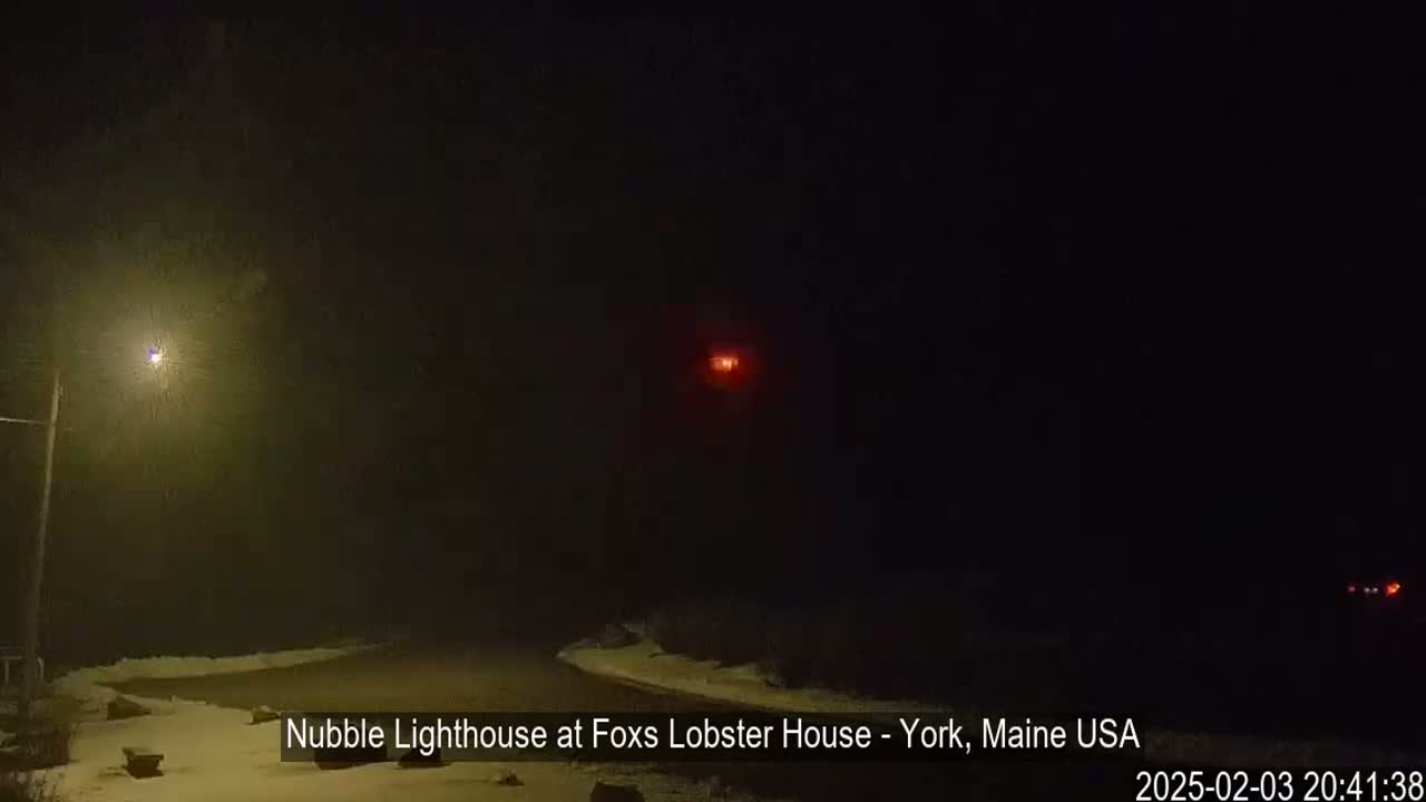 A snow-covered road curves into a dark, foggy night, illuminated by a streetlight and a distant red light.