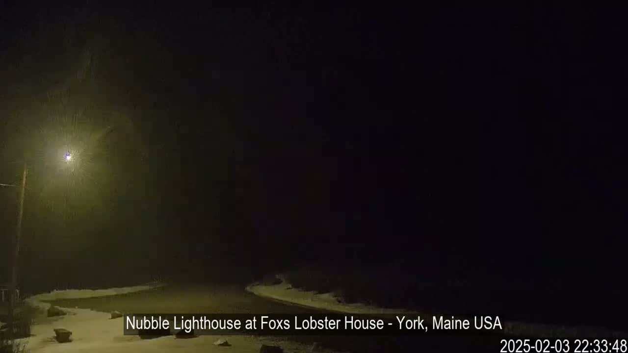 A snow-covered road curves gently into a dark, snowy night scene illuminated by a single streetlight.
