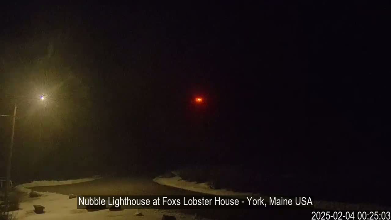 A nighttime scene showing a snow-covered roadway leading towards a distant, lit lighthouse under falling snow.