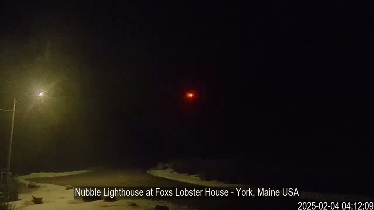 A dimly lit snowy scene at night shows a distant red light and a streetlight illuminating a small area of a road.