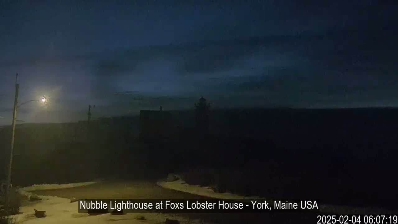 A dimly lit lighthouse is visible in the distance under a dark, mostly cloudy night sky, with a snowy foreground.