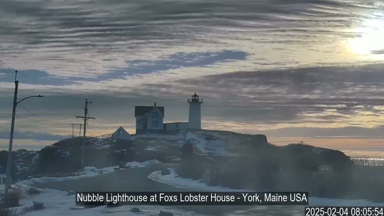 A lighthouse and adjacent buildings are situated on a snow-covered, rocky coast under a mostly cloudy, sunrise sky.
