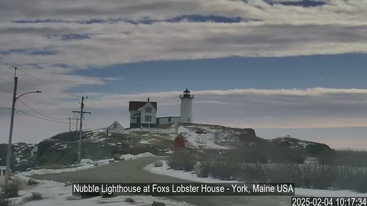 A lighthouse and a house sit atop a snow-dusted hill under a mostly cloudy sky.