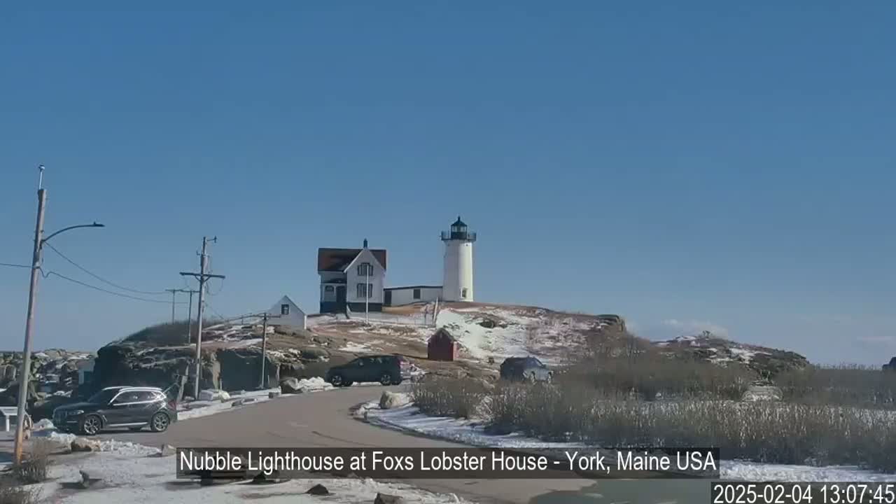 A white lighthouse stands on a snow-dusted hilltop overlooking a road with parked cars under a clear blue sky.