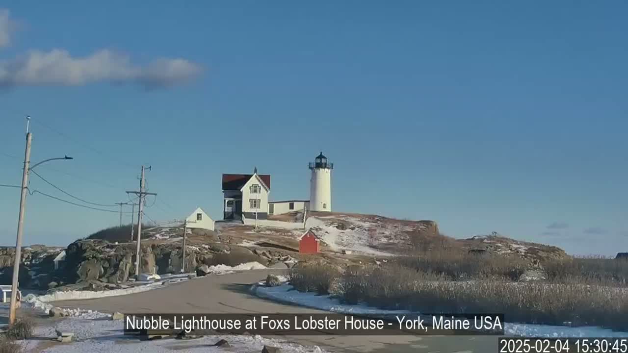 A white lighthouse and adjacent buildings stand on a snow-dusted, rocky hill under a mostly clear, sunny sky.