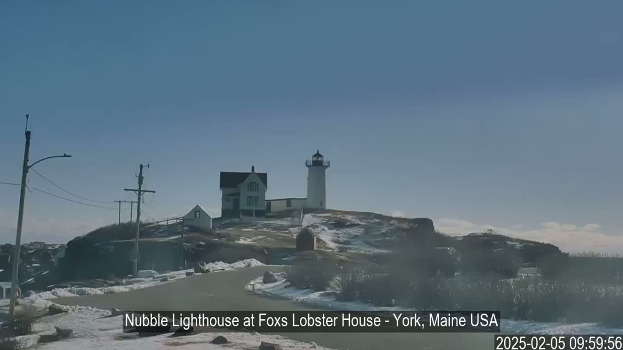 A lighthouse and adjacent buildings sit atop a snow-dusted hill on a clear, sunny day.
