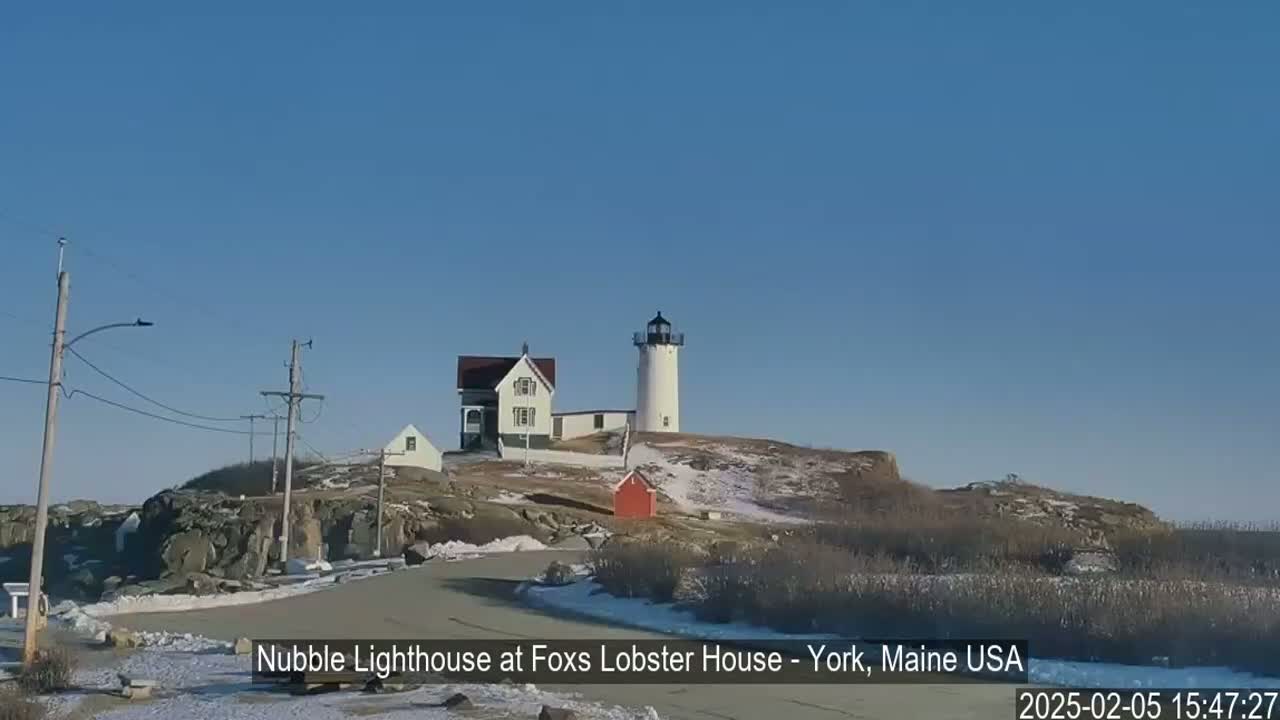 A white lighthouse stands on a rocky hill, partially covered in snow, next to a smaller house, under a clear blue sky.