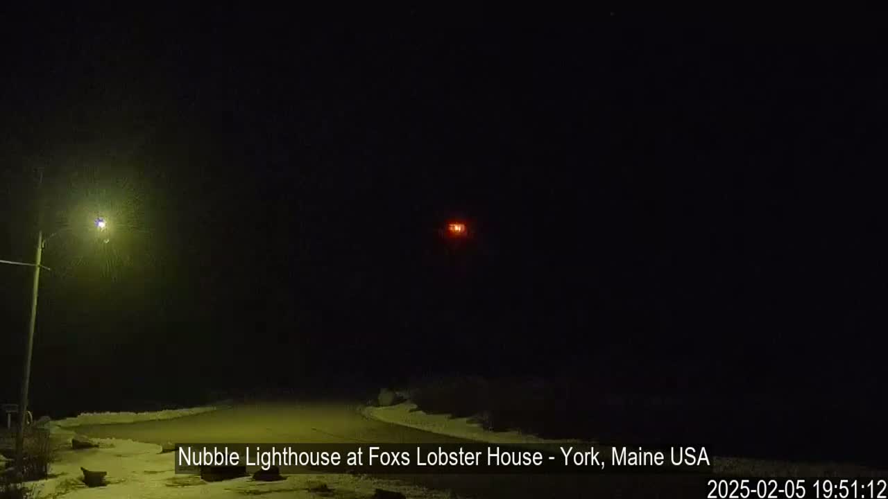 A dark, snowy nighttime scene shows a lit-up lighthouse in the distance, illuminated by a streetlight closer to the viewer.