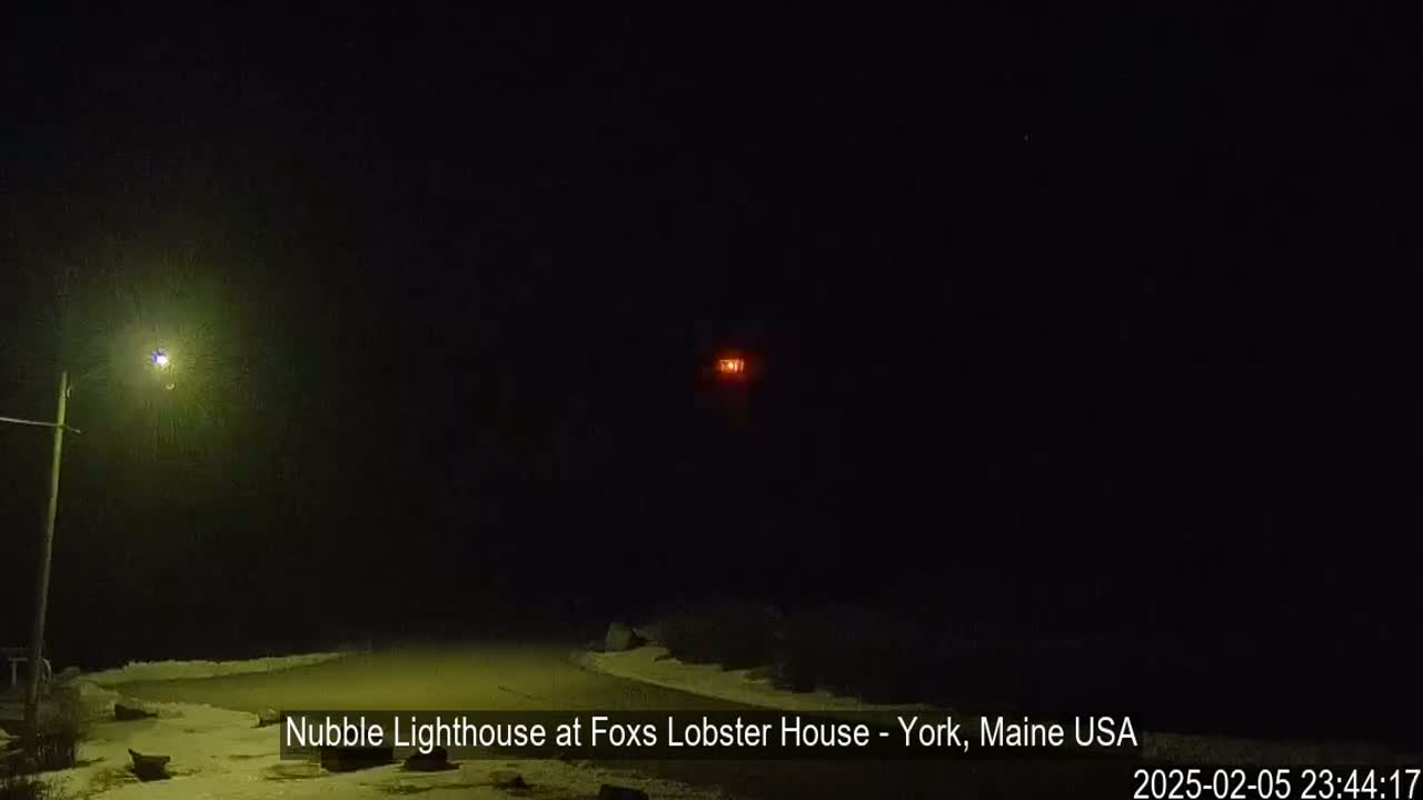 A nighttime view of a snow-covered shoreline with a distant, lit lighthouse and a streetlight illuminating a small area.