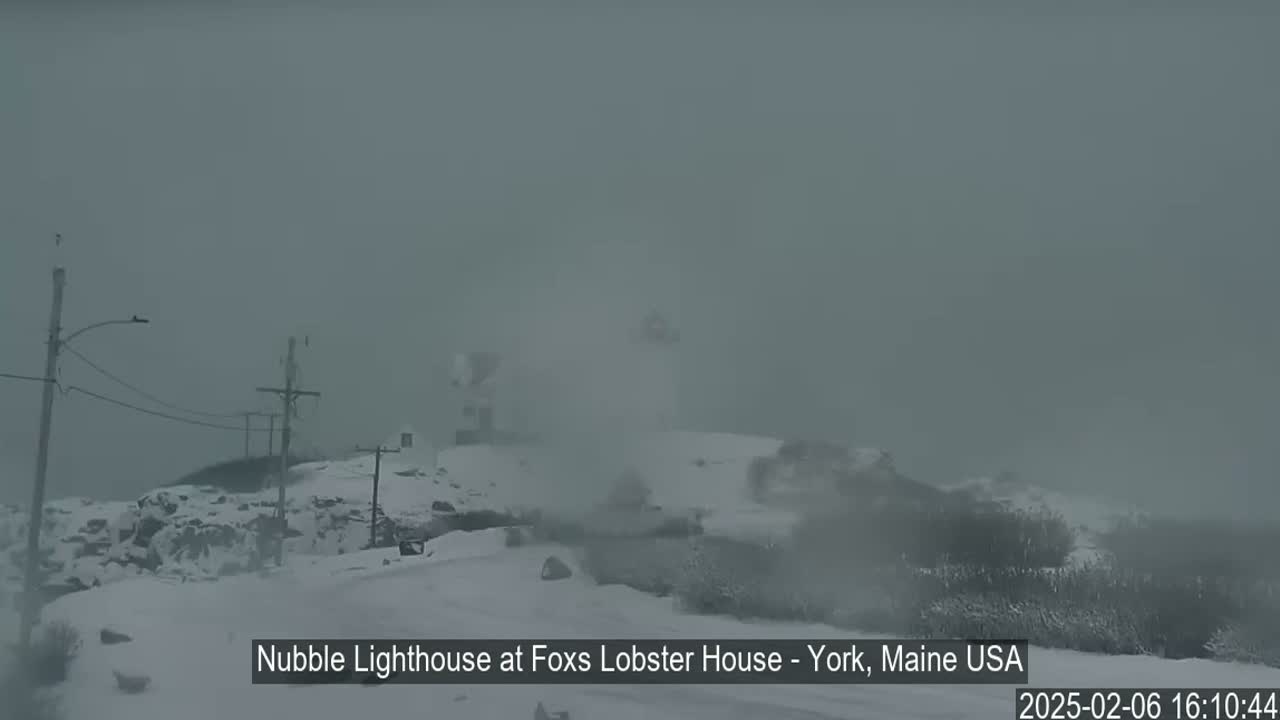 A snow-covered landscape features a lighthouse in the distance, partially obscured by heavy fog or blowing snow.