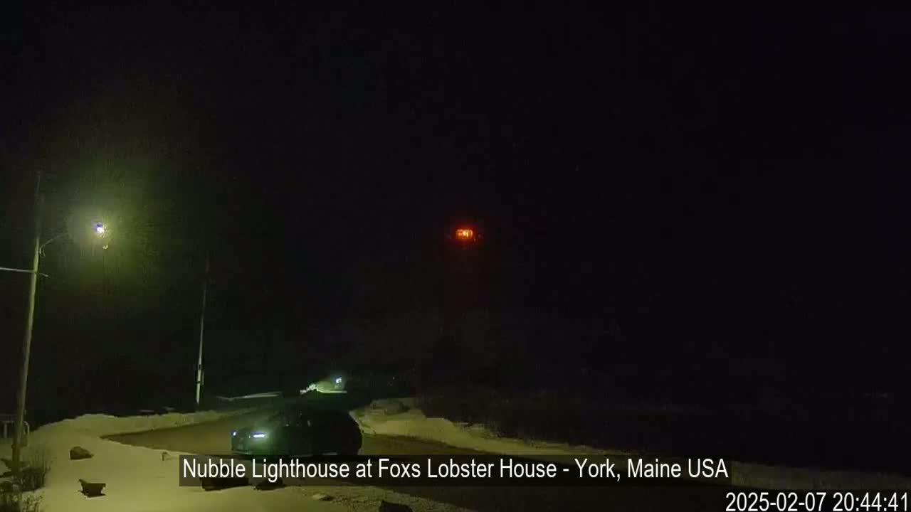 A nighttime view shows a snow-covered landscape with a vehicle parked near a lit-up lighthouse in the distance under a dark sky.