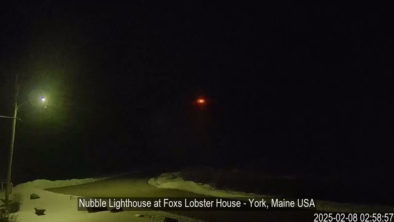 A nighttime scene shows a lighthouse's light in the distance, a streetlight nearby illuminating a snow-covered area, under a dark, hazy sky.