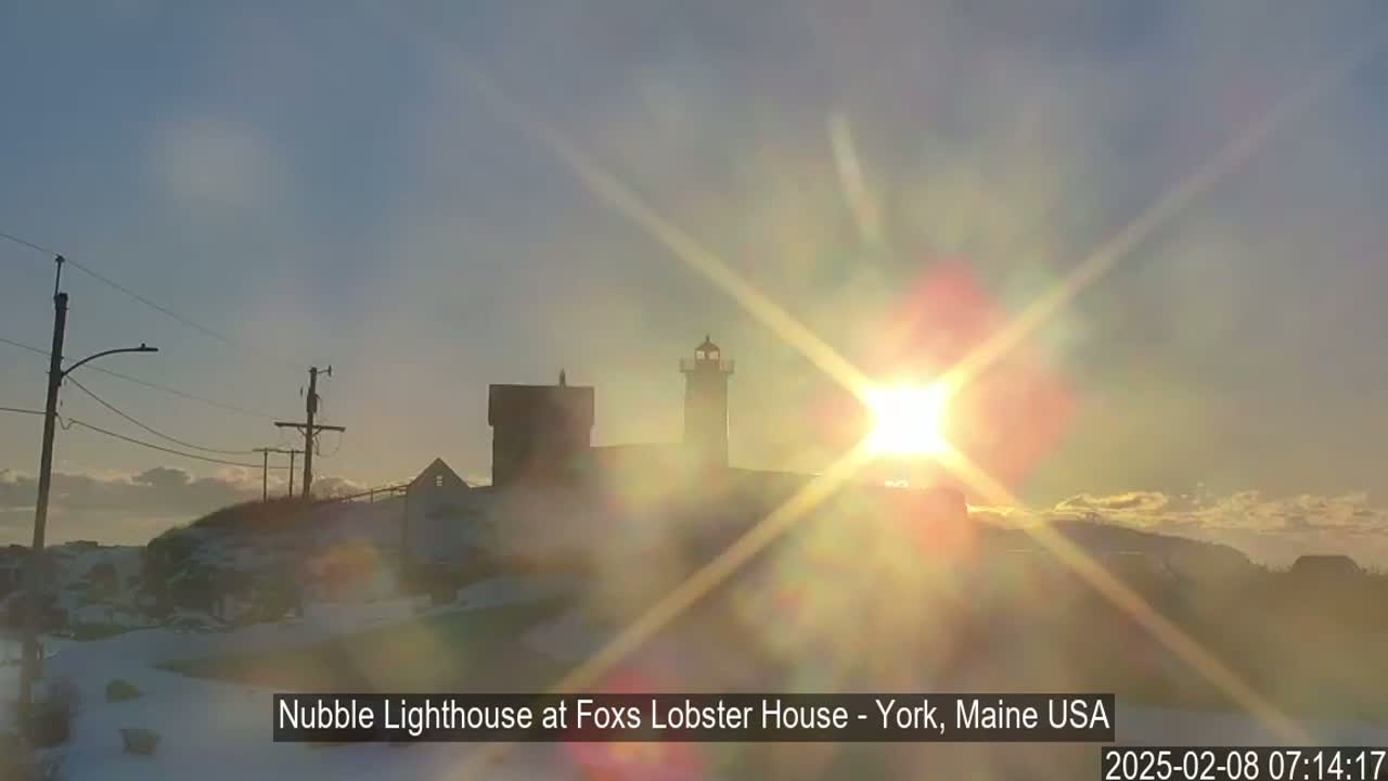 A lighthouse and other buildings are silhouetted against a bright, sun-filled sky, with a light dusting of snow on the ground.