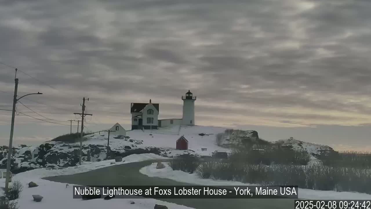 A lighthouse and other buildings sit on a snow-covered, rocky hill under an overcast sky.