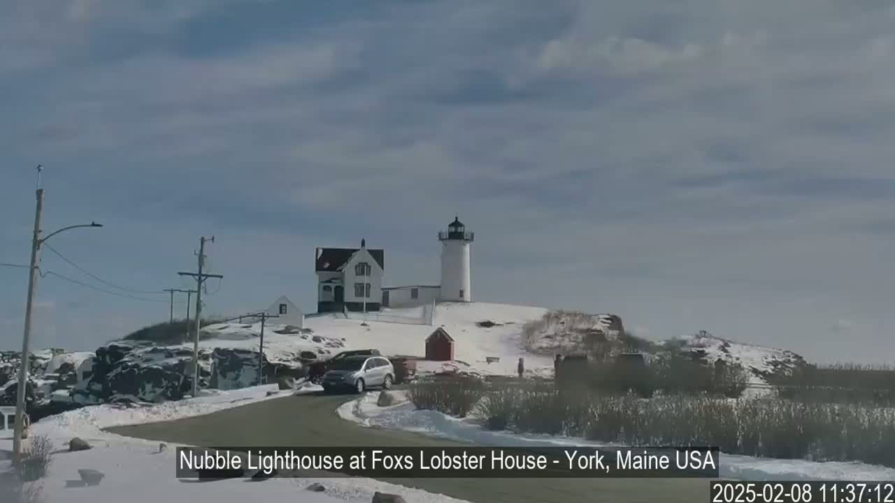 A lighthouse and adjacent building sit atop a snow-covered hill, with a car parked on a road leading to them under a partly cloudy sky.