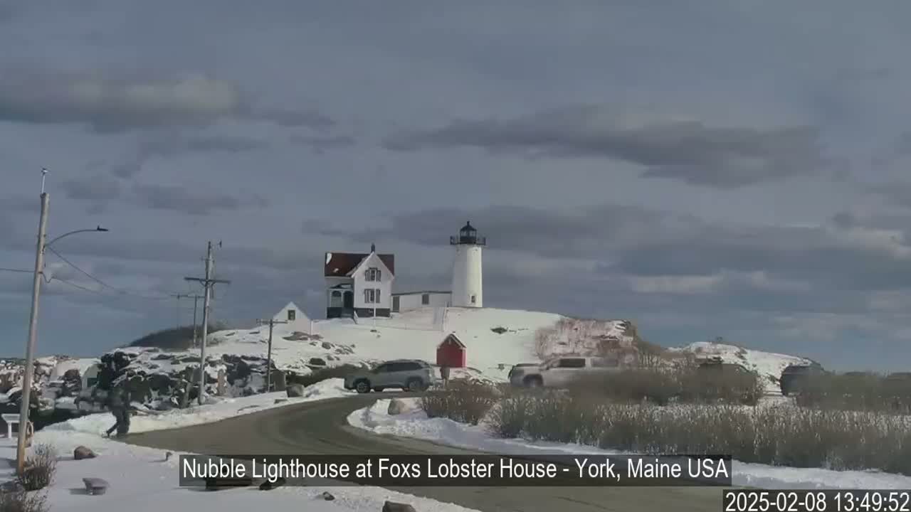 A white lighthouse sits atop a snow-covered hill next to a house and some vehicles, under a mostly cloudy sky.