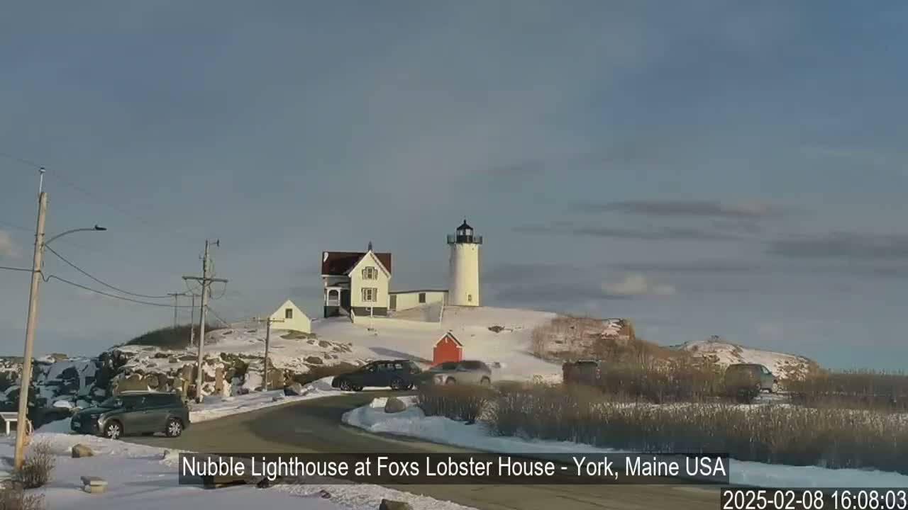A lighthouse and nearby buildings are situated on a snow-covered hill overlooking a road with several cars parked, under a mostly clear sky.