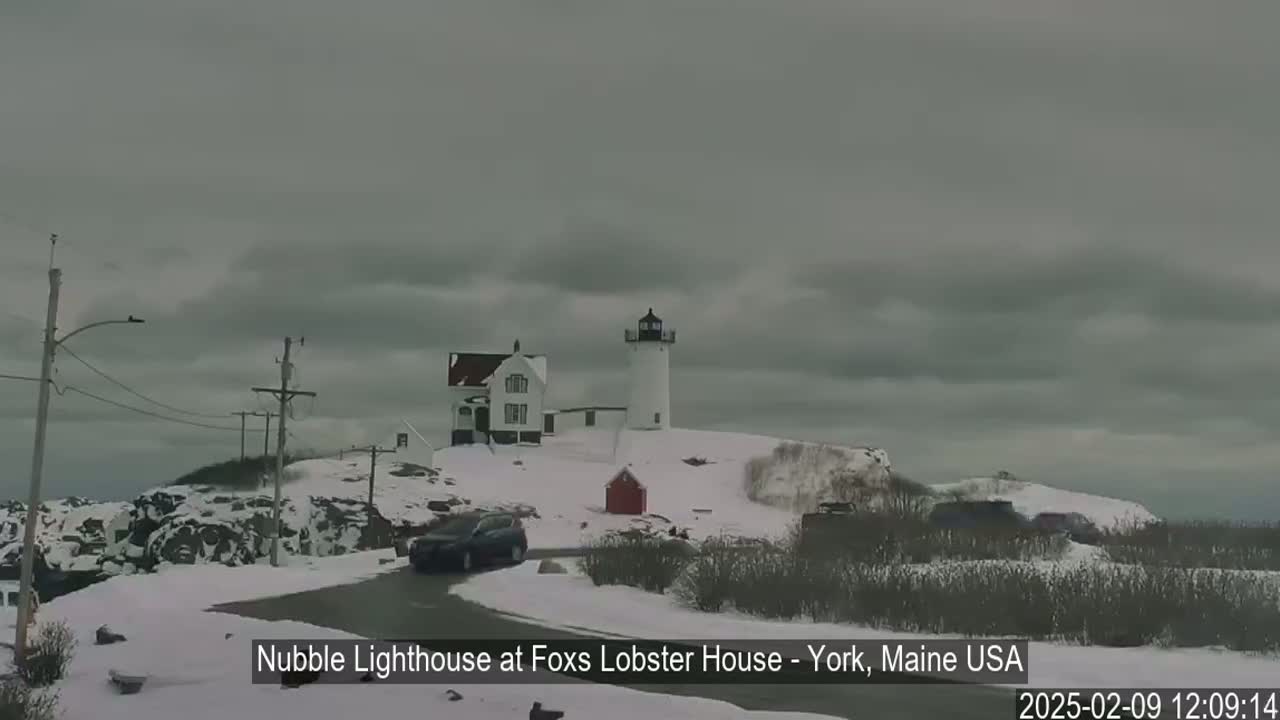 A lighthouse and a house sit atop a snow-covered hill under an overcast sky.