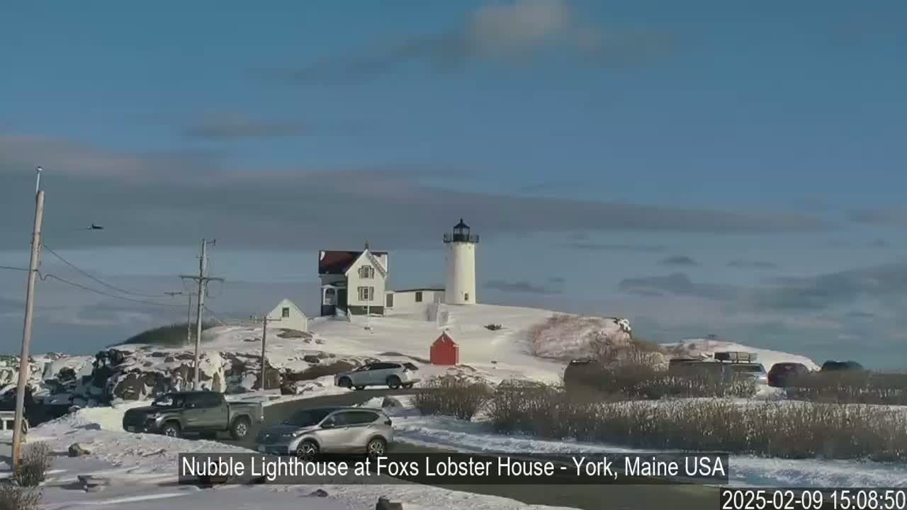 A lighthouse and several cars are situated on a snow-covered hill overlooking a partially-cloudy sky.