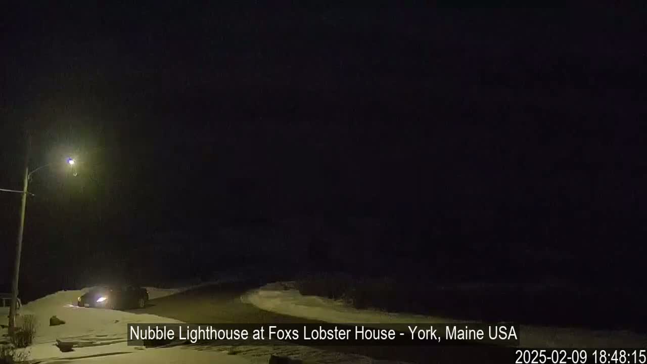 A dark nighttime scene shows a snow-covered road with a vehicle parked near a lit streetlamp.