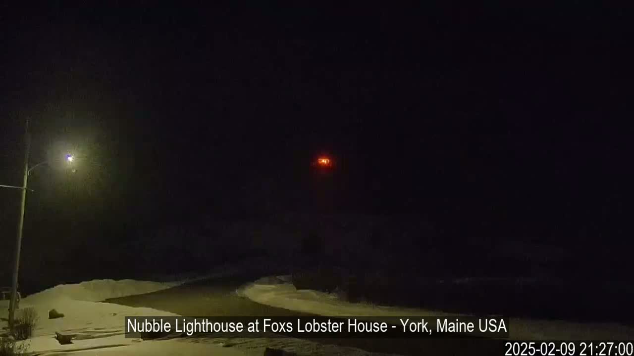 A nighttime, snowy scene shows a lighthouse with a lit lamp in the distance, illuminated by a streetlight closer to the viewer.
