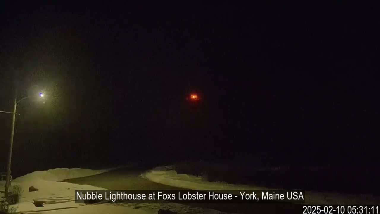 A snow-covered shoreline is visible at night, illuminated by a streetlight and the distant, faint red glow of a lighthouse.