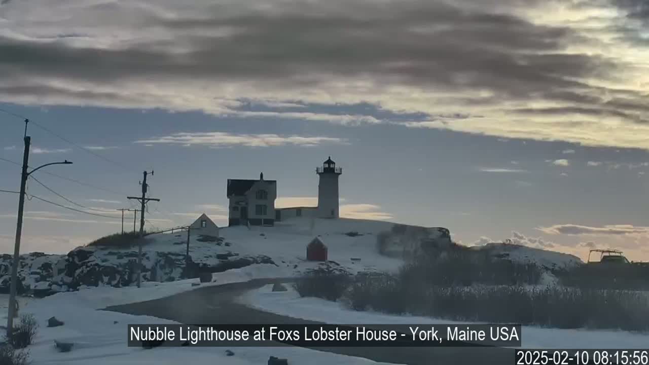 A snow-covered lighthouse and adjacent buildings sit atop a rocky outcrop under a partly cloudy sky.