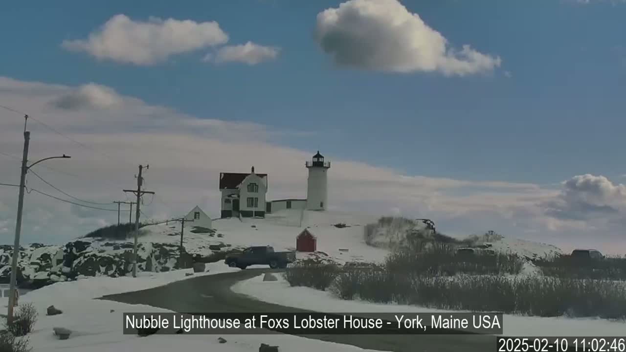 A lighthouse and accompanying buildings sit on a snow-covered rocky outcrop under a partly cloudy sky.