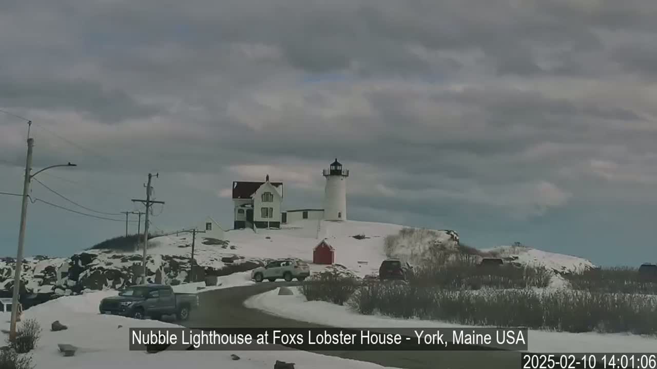 A lighthouse and accompanying building sit atop a snow-covered hill under a cloudy sky, with several vehicles parked nearby on a snow-covered road.