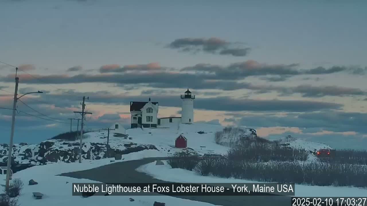 A lighthouse and adjacent building sit on a snow-covered hill under a mostly cloudy, twilight sky.