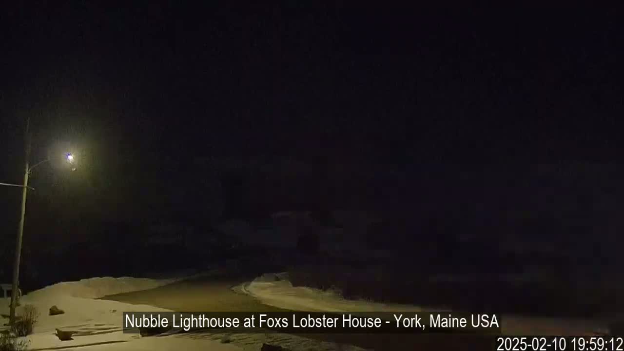 A nighttime scene shows a snow-covered area illuminated by a single streetlight, with dark buildings and a dark sky.
