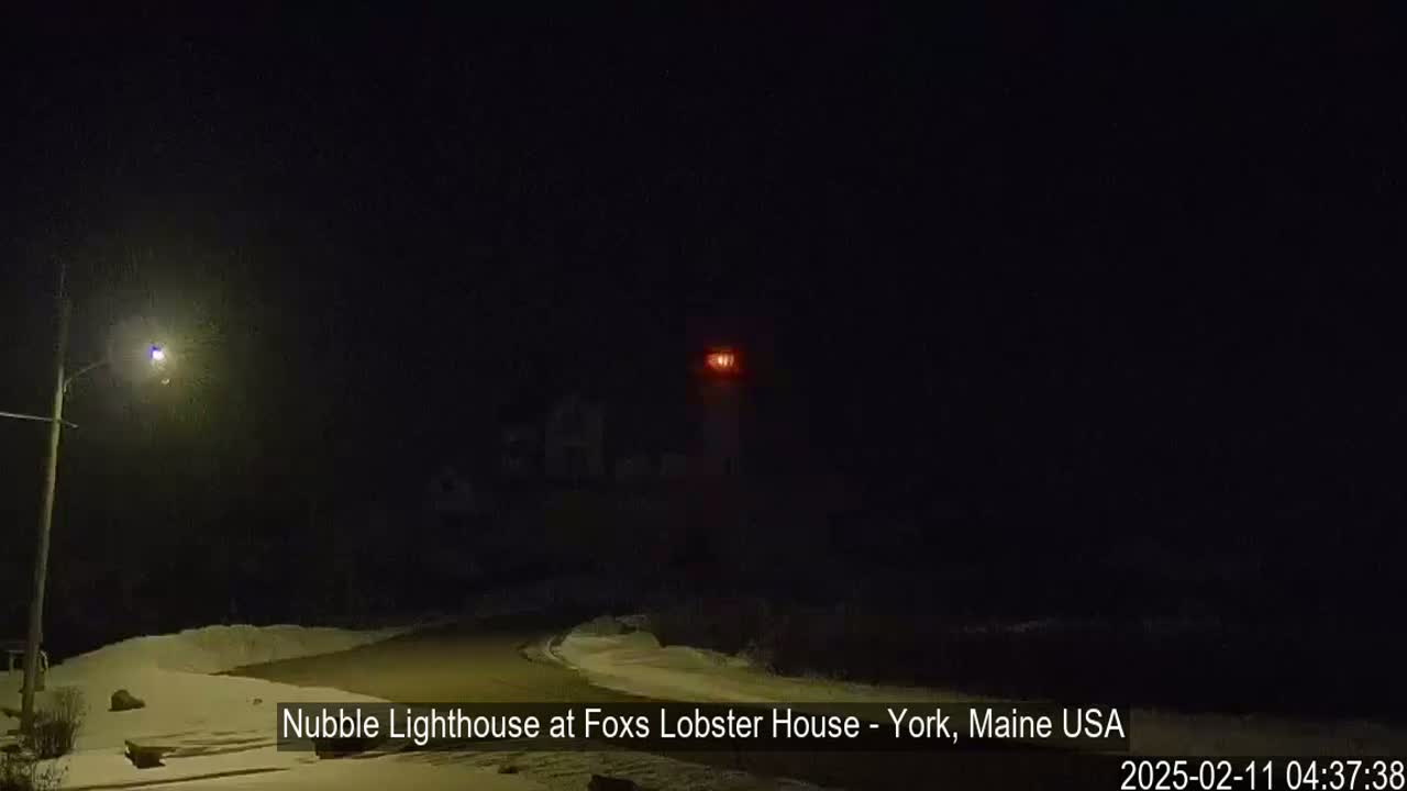 A nighttime view of a lighthouse emitting a red light, illuminated by a streetlamp, in a snow-covered landscape.