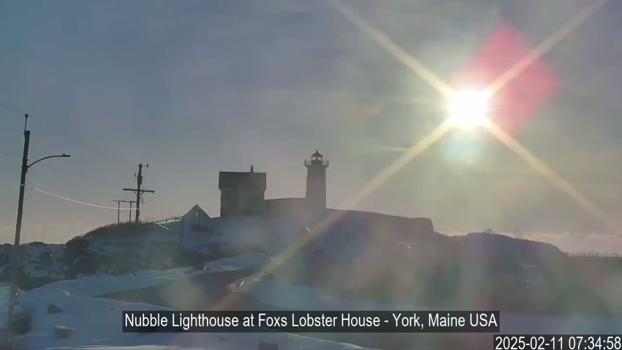 A lighthouse and other buildings sit on a snow-covered hill, silhouetted against a bright sun with lens flare.